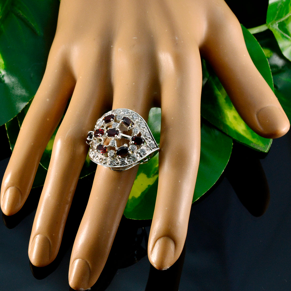 Rosa abstract red stone ring with silver setting displayed on a mannequin hand against green leaves