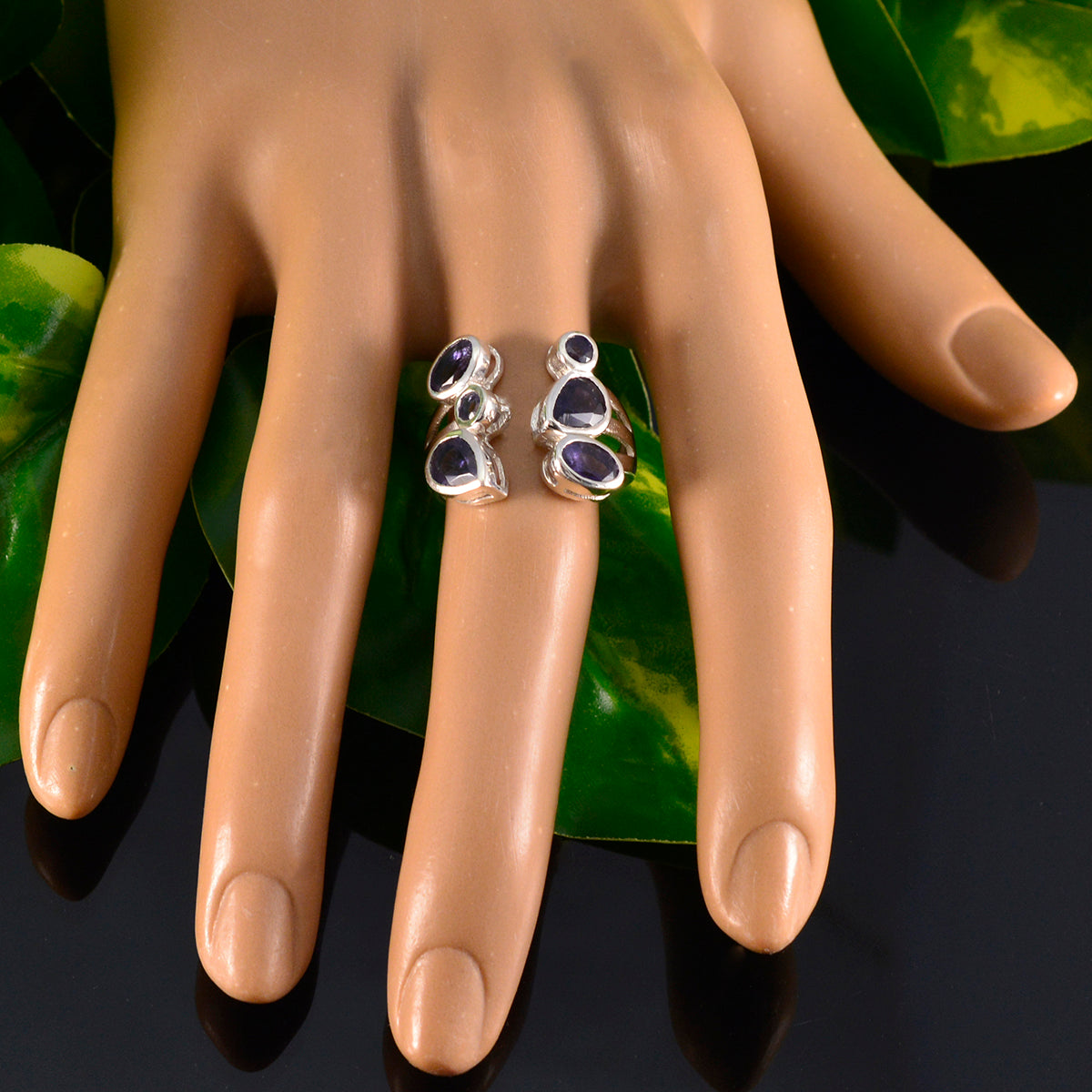 Genesis blue gemstone statement ring with silver setting worn on mannequin hand over green leaves background