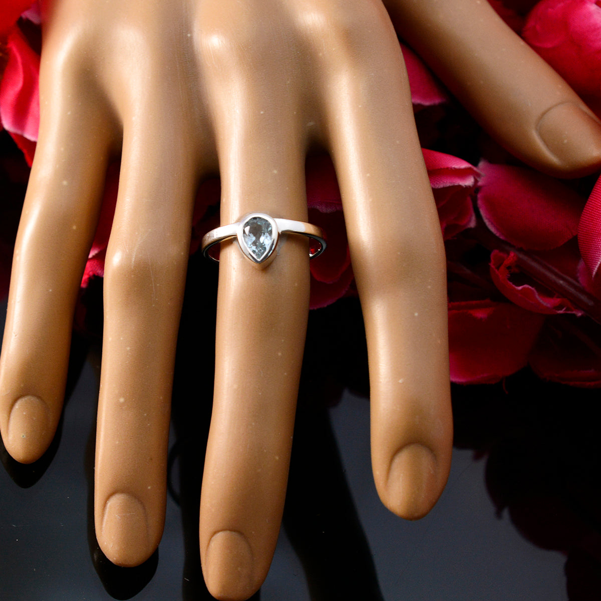 Natcha blue solitaire ring with pear-shaped blue gemstone on display hand with red petals background