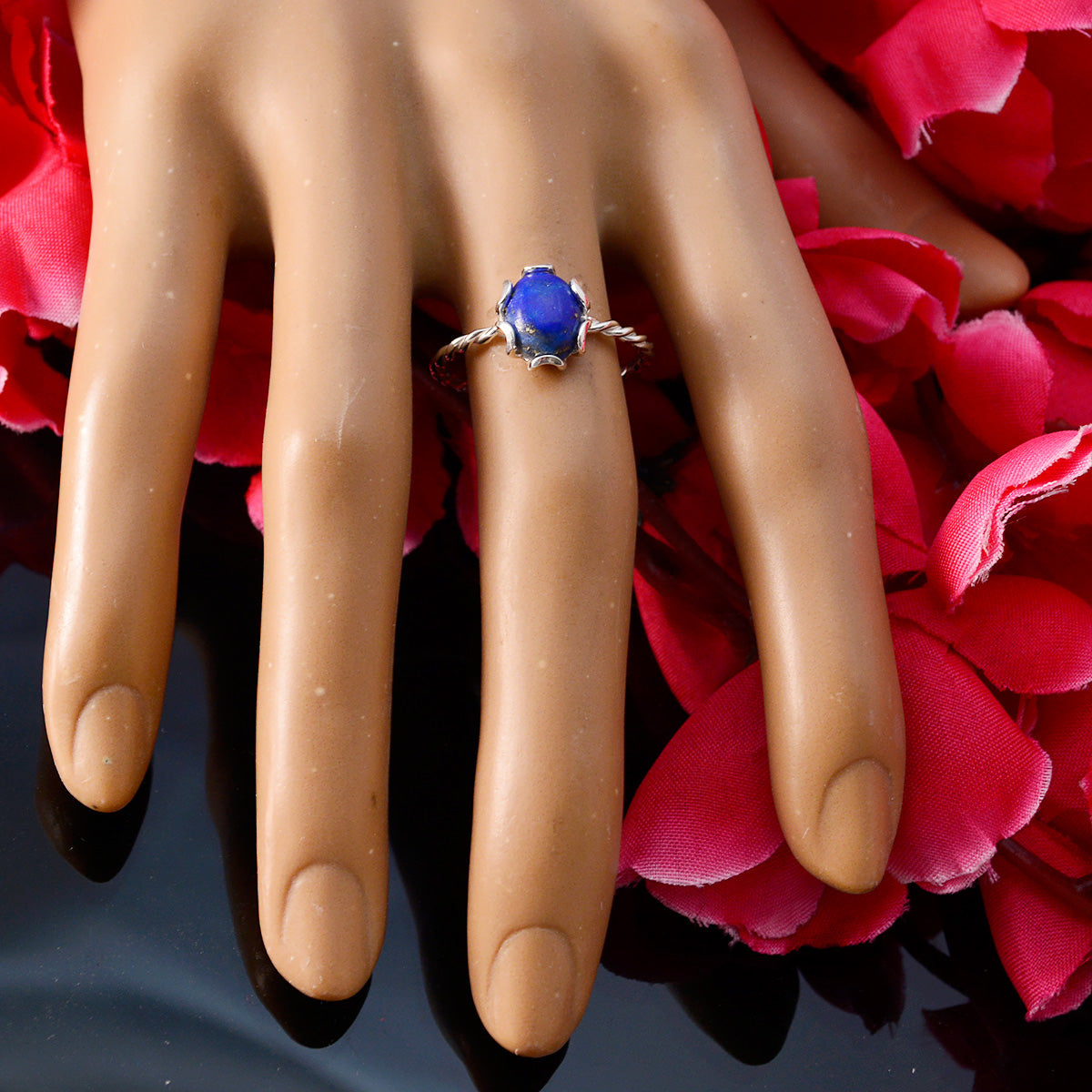 Riya blue solitaire twist ring with a vibrant blue stone on a display hand surrounded by red petals