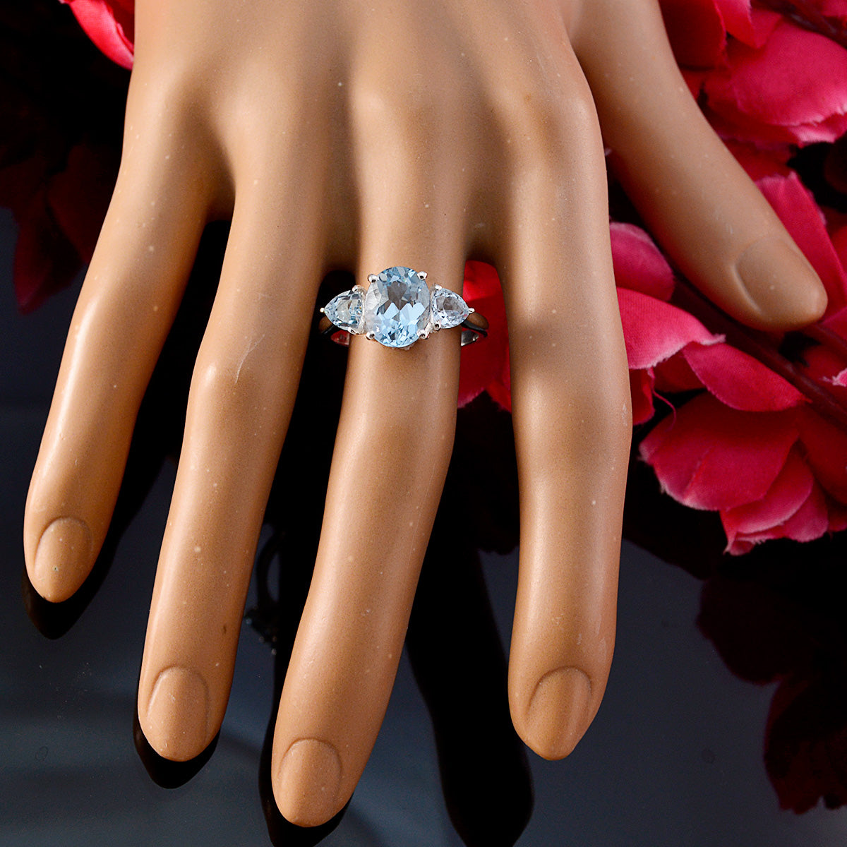 Chloe blue gemstone trilogy ring with three blue stones on a hand display against red petals